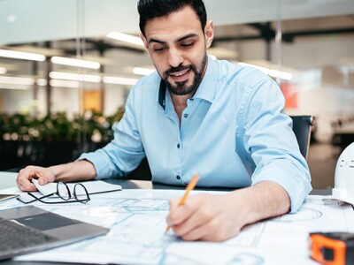 Engineer working on technical drawings with a laptop and safety helmet nearby.