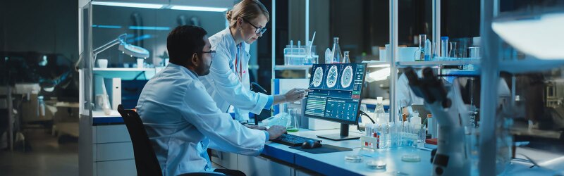 Two scientists in lab coats examine brain scans on a computer screen in a lab filled with equipment and beakers.