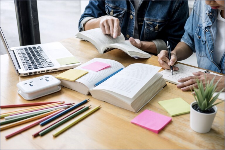 Two students working at table with book, laptop, Post-it notes, and colored pencils
