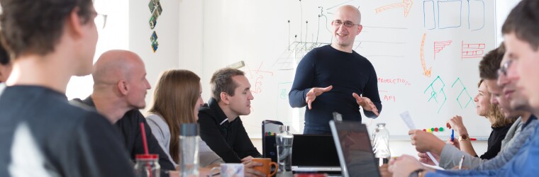 Man stands in front of tech infrastructure plan on whiteboard while in a meeting with coworkers