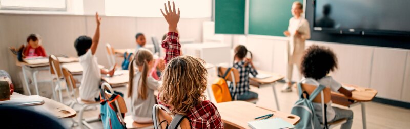 A classroom of students raise their hand to answer female teacher.