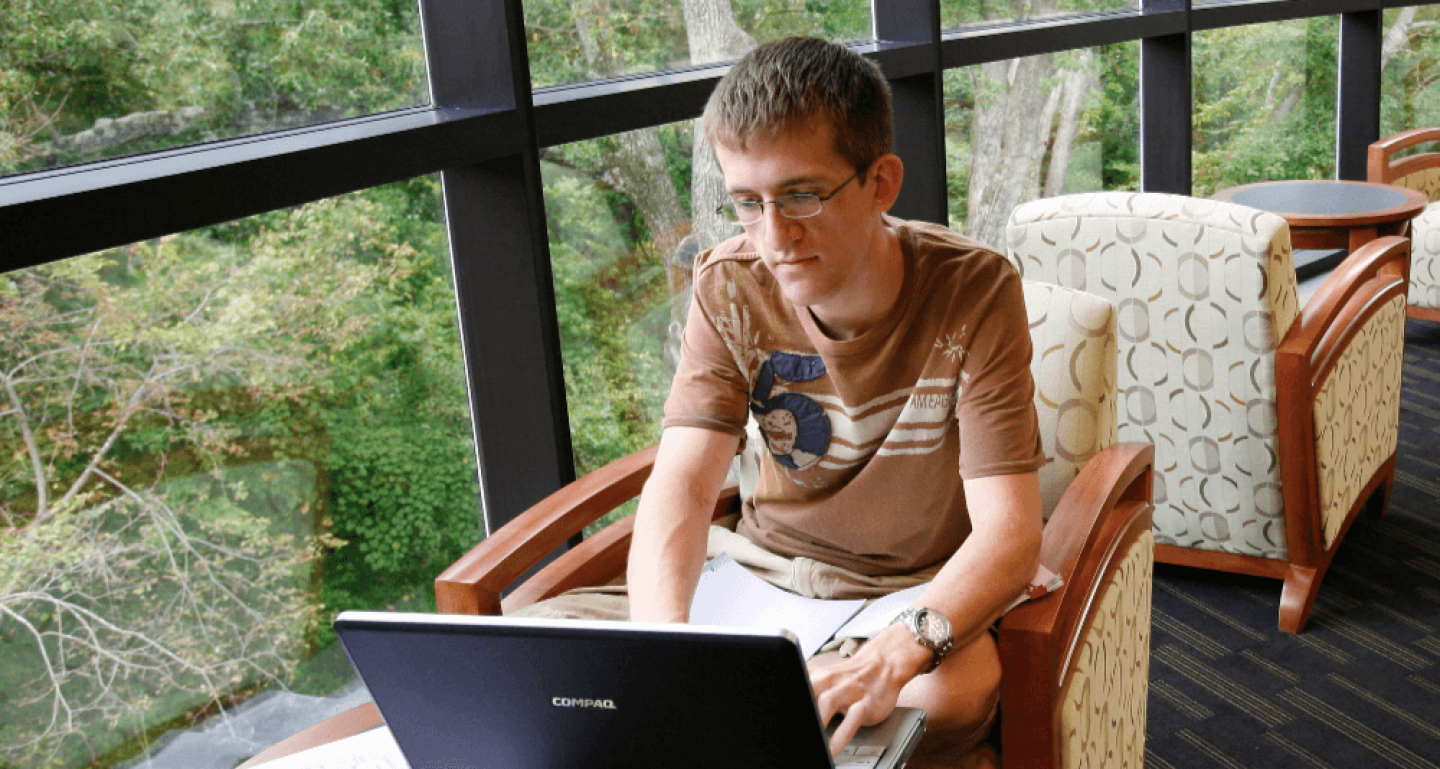 William & Mary student seated in front of window working on laptop next to textbook