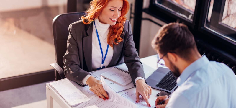 A female investment banker sits at a desk advising a male client where to sign on a document.