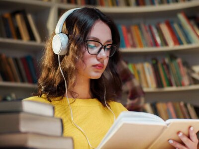 Young woman in glasses and headphones studying for her PHD, reading a book and listening to music in the library.
