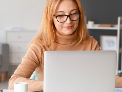 Seated at a laptop in an office, a woman wearing glasses takes notes as she types.
