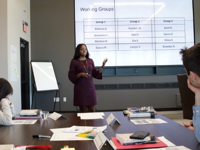 Woman speaking at front of room pointing at chart projection