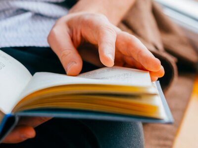 Young man reading a book in his lap while sitting next to a window.