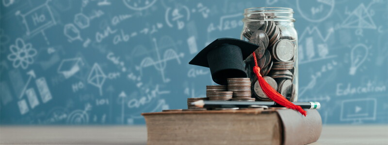 A tiny graduation cap, stacks of coins and a glass jar filled with coins are stacked up on a book.