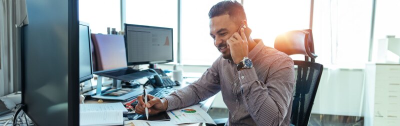 Male financial analyst discussing financial reports with colleague over mobile phone sitting at his office desk.