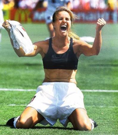 Soccer player Brandi Chastain raising arms and cheering while kneeling on field