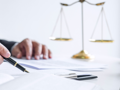 Hand holding fountain pen over business documents. Scales of justice is in the background, while a gavel is seen in the foreground.