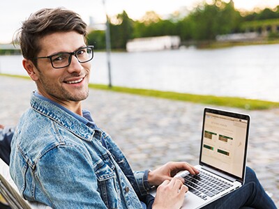 Man-In-Jean-Jacket-With-Glasses-And-Laptop-Sitting-On-Bench-In-Front-Of-Path-And-Water