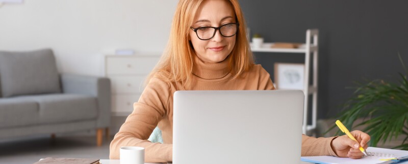 Seated at a laptop in an office, a woman wearing glasses takes notes as she types.