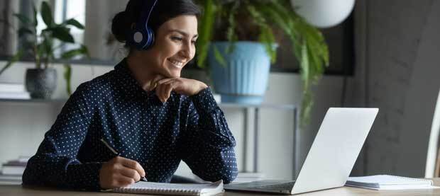 Smiling student wearing headphones and taking notes while seated at a laptop