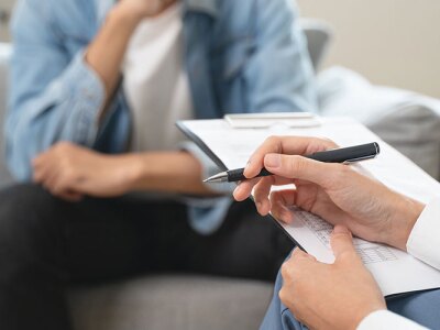 A counselor, seen from behind, holds a pen and clipboard while a client sits on a sofa.