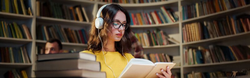 Young woman in glasses and headphones reading a book and listening to music in the library.