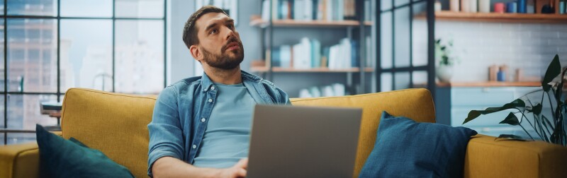 Man sitting on a sofa with a laptop, deep in thought, reflecting on possibilities