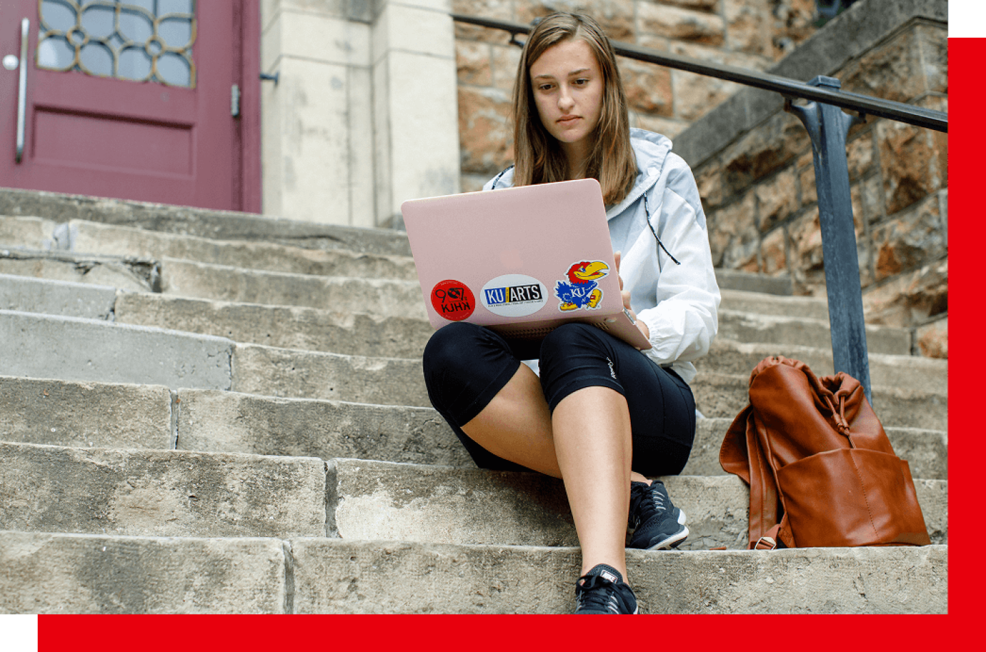 Woman working on laptop with KU stickers while sitting on steps outside