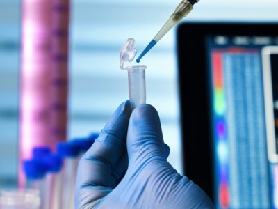 Gloved hands in a lab transfer liquid from a dropper to a vial.