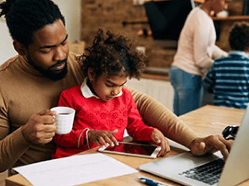 Man with child on lap holding coffee and using laptop