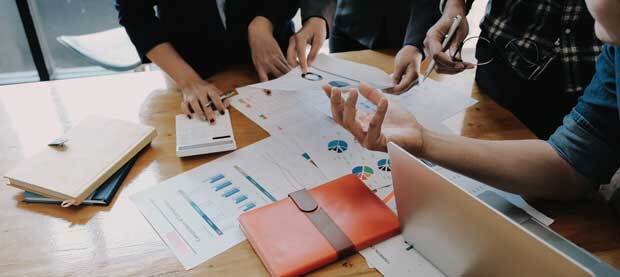 Four people in discussion around a table that's piled with papers, books and a laptop