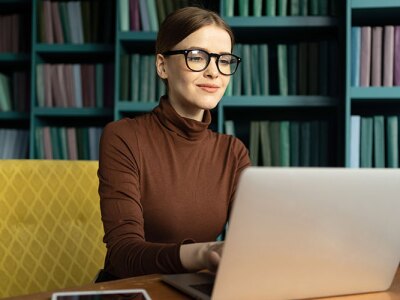 A woman in glasses studying on a laptop with bookshelves in the background, representing an online education program for teaching.