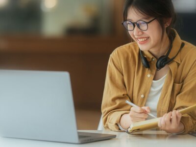 Asian girl student reviews her college application status on her laptop while taking notes.