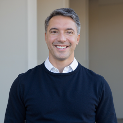 Headshot of Jason Vanecko smiling, wearing a black sweater over a white collared shirt, with a softly blurred neutral background