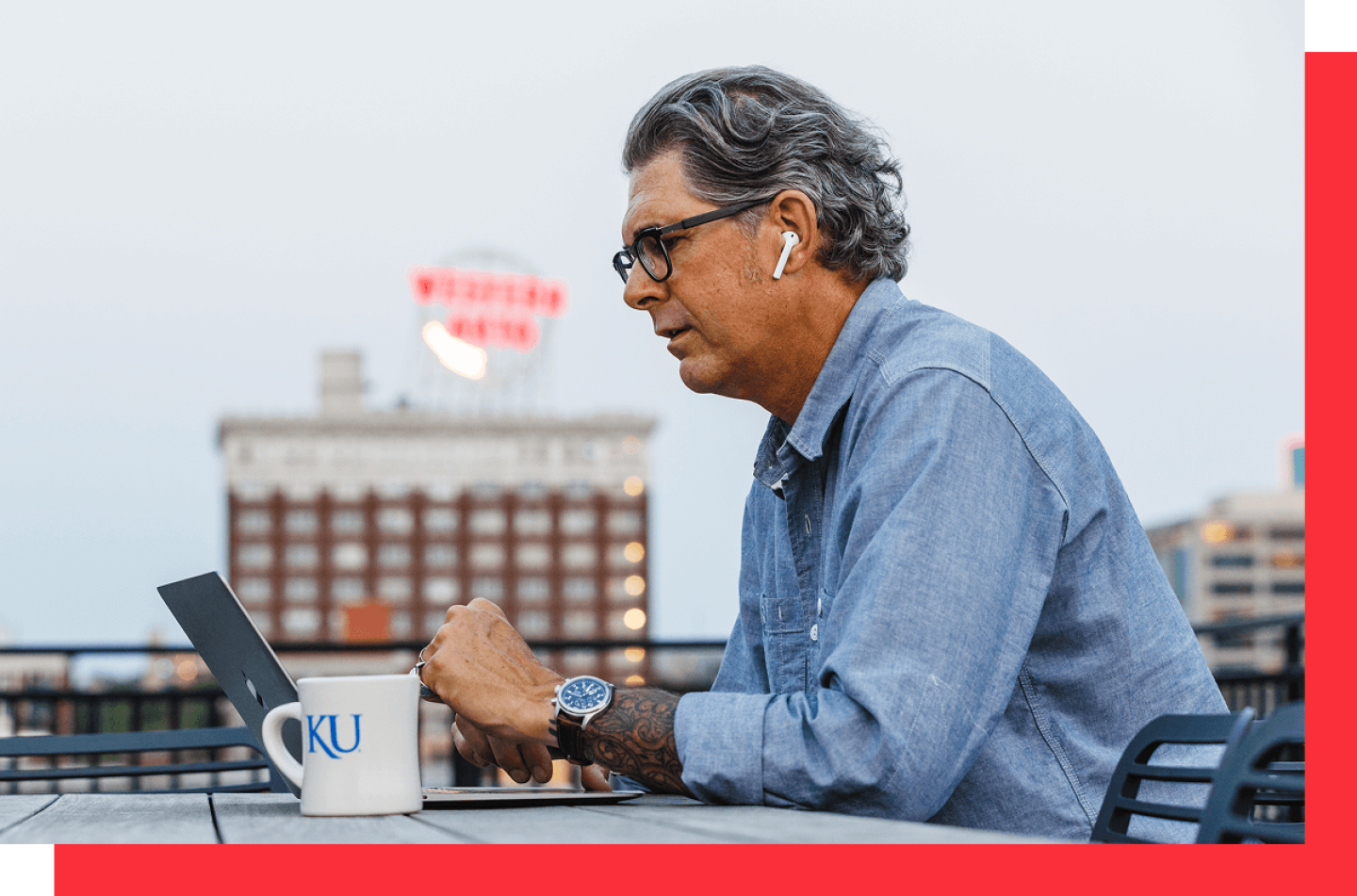 Man in glasses working on a laptop outdoors with a KU coffee mug