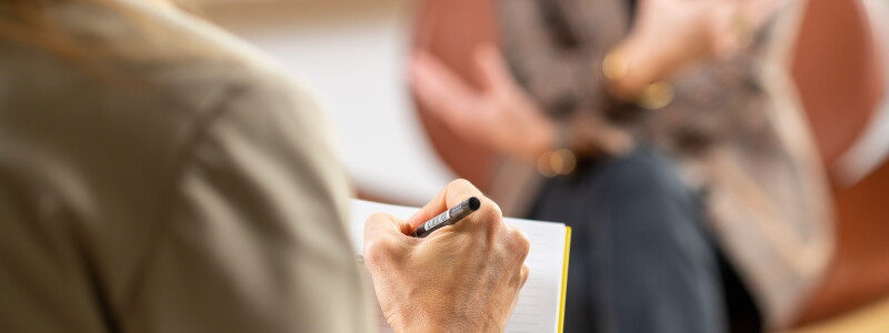 Looking closely over the shoulder of a counselor who takes notes while a woman sits across from them speaking.