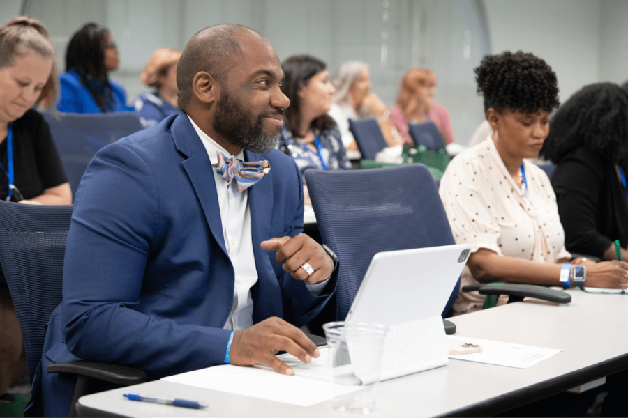 Man in blue suit with bow tie listening to speaker and using laptop