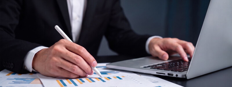 A person in a suit reviews financial charts and bar graphs with a stylus while typing on a laptop.