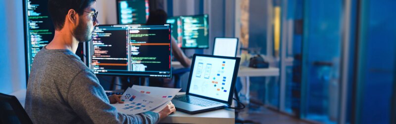 Man wearing glasses sits in front of multiple computer screens. He is holding a print out of the diagram shown on the laptop screen.