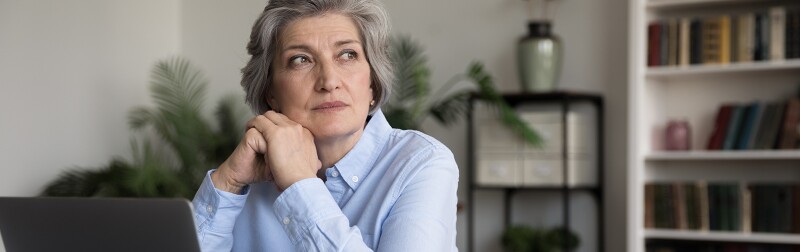 Thoughtful serious mature grey haired business lady sitting at work office table with laptop
