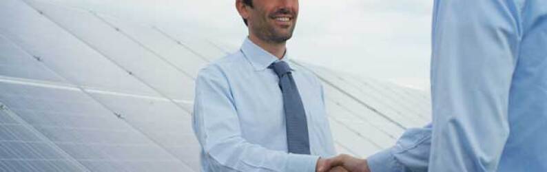 Two businessmen, one in a hard-hat, shake hands in front of a bank of solar panels.