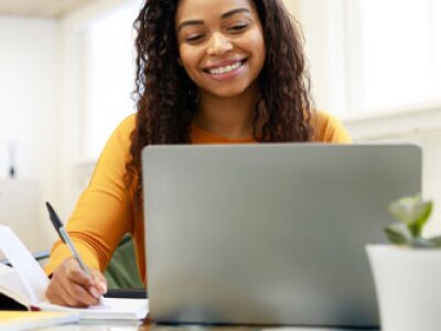 Woman sitting at desk, using computer and writing in notebook
