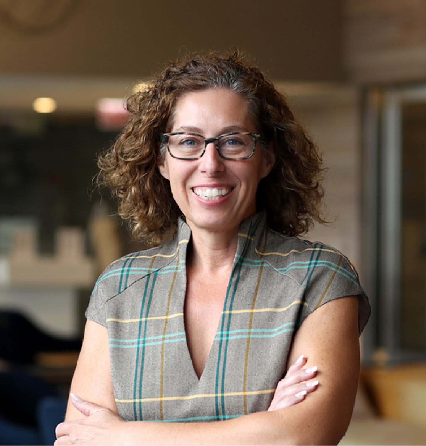 Headshot of Teelina House with curly hair and glasses, arms crossed, wearing a plaid top in a softly lit indoor setting.