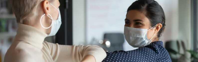 Two office workers wearing facial masks bump elbows in greeting.