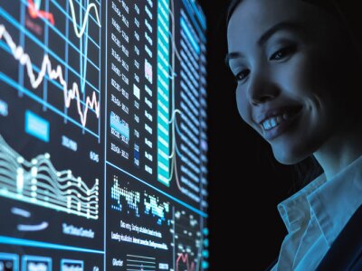 Young female data analyst reviews analytic metrics on touch screen wall monitor.