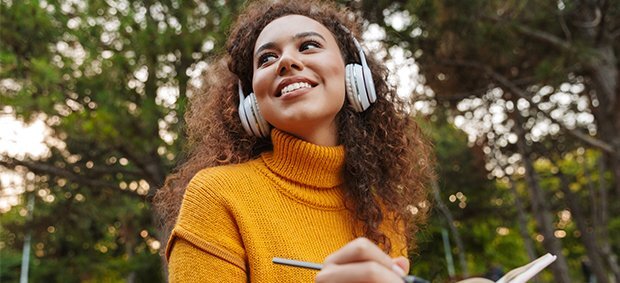 woman-in-yellow-turtleneck-wearing-headphones-writing-in-notebook-in-front-of-tree