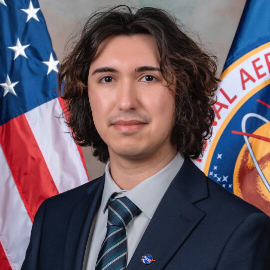Rudy Ruano standing in front of flags from the USA and NASA