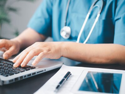 A nurse in scrubs typing on a laptop with a stethoscope around their neck, medical documents, and a tablet on the desk.