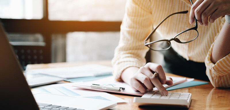 A woman sitting at a desk, holding a pair of glasses and typing on a calculator.