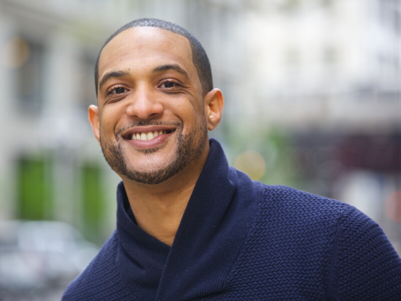 Man in a navy blue sweater smiling at the camera with a cityscape in the background