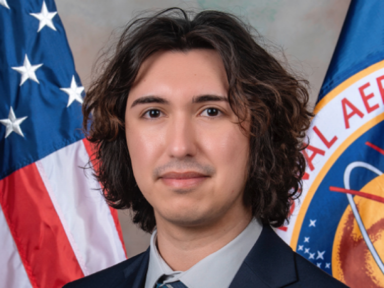 Rudy Ruano standing in front of flags from the USA and NASA