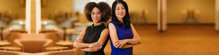 Two smiling female students stand back to back with their arms crossed