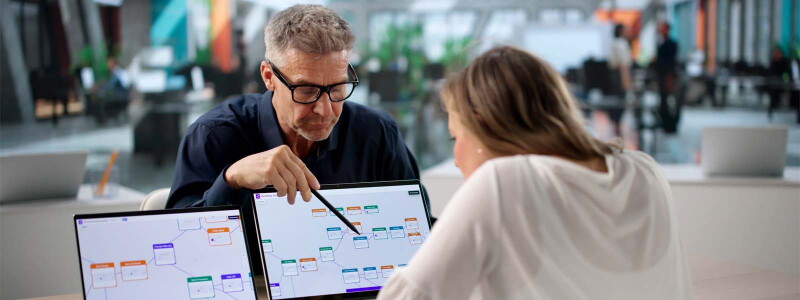 Two business professionals at a desk looking at a workflow spread across two computer screens.