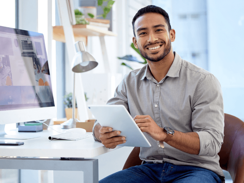 Man seated at desk holding a tablet and smiling in front of desktop monitor