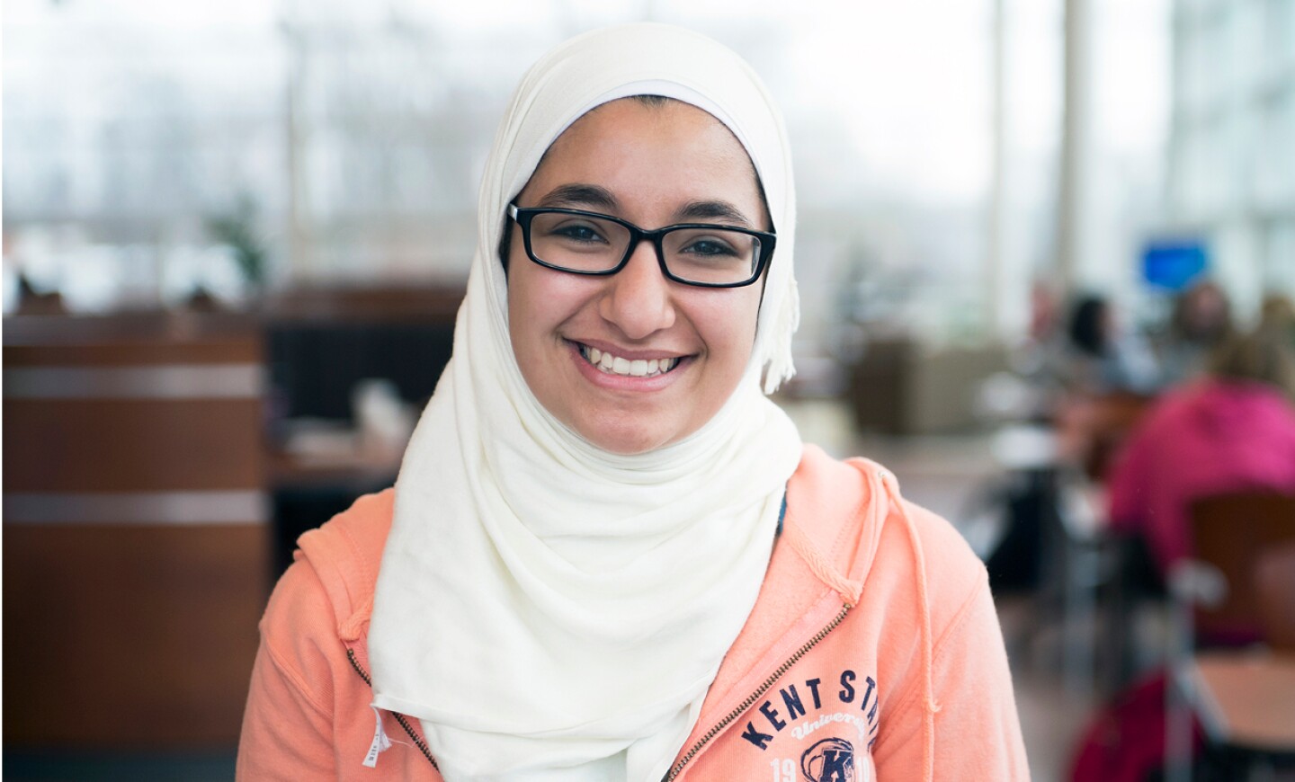 Smiling student wearing glasses and a white headscarf inside a campus study area