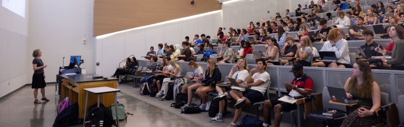 Wide shot of a female KU professor standing in front of a class in a lecture hall on campus.